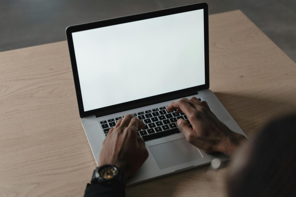 A person typing on a laptop with a blank screen on a wooden desk, ideal for mockups.